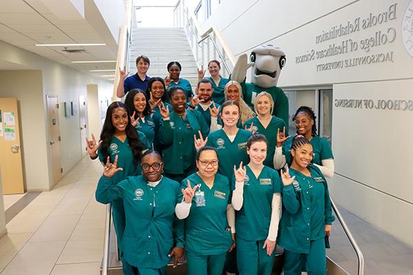 Cohort of ABSN students pose with Dunk'n the Dolphin on the steps of the Keigwin School of Nursing staircase.